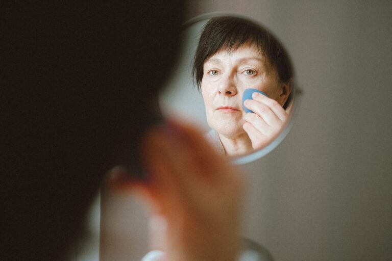 Elderly woman looking at herself in the mirror while applying makeup sponge.
