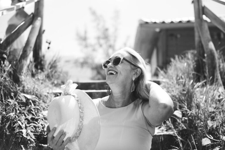 A smiling senior woman enjoys a sunny day outdoors wearing sunglasses and a sunhat.